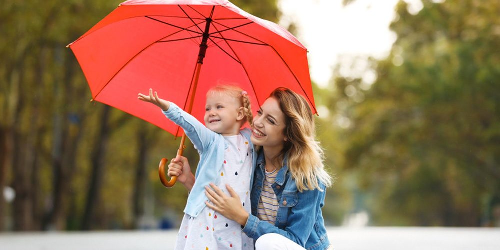 Adult kneeling beside child holding a red umbrella on a tree-lined path - https://www.rogersvilleins.com/